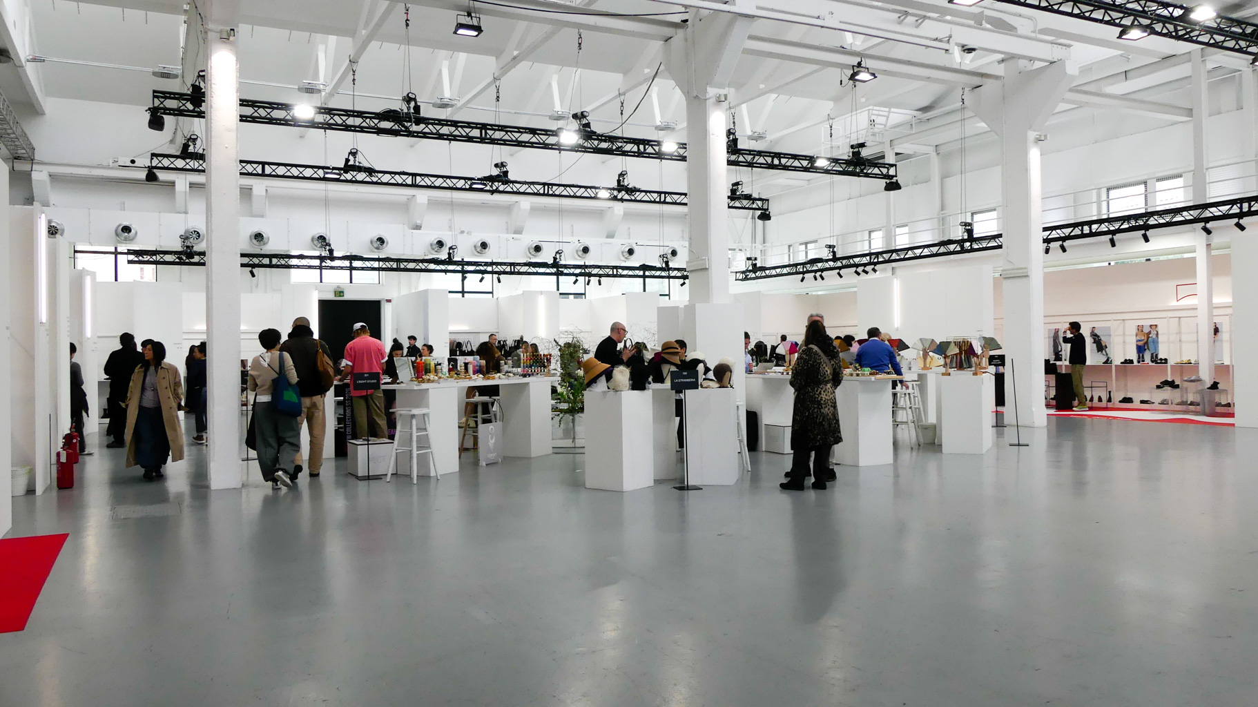 Spacious white exhibition hall with high ceilings and industrial lighting rigs, filled with visitors browsing white display tables and booths at a fashion or trade show event.